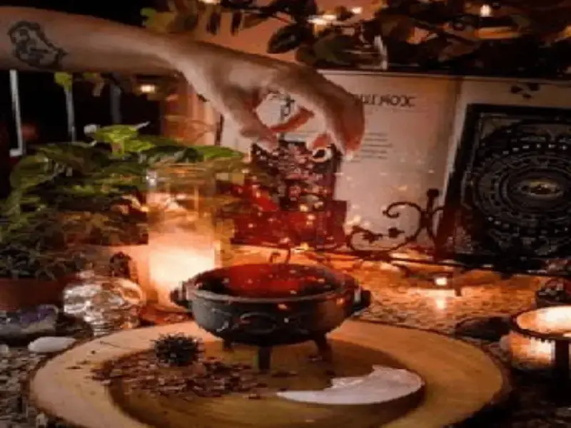 Mano agregando ingrediente mágico a un pequeño caldero, rodeado de velas encendidas, plantas, libro de fórmulas abierto, luna y semillas, representando las conexiones del destino al contactarnos.
