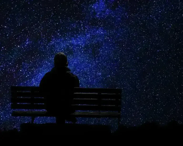 Silueta de un hombre sentado en una banca de madera bajo un cielo nocturno estrellado, representando el momento introspectivo de hablar con el universo y manifestar deseos.
